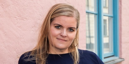 Person med långt blont hår i mörkblå tröja står lutad mot en rosa vägg vid ett blått fönster.