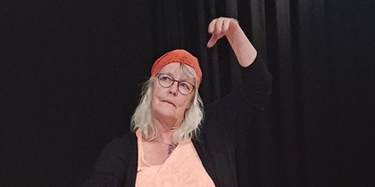 Kvinna med glasögon och röd sjal på huvudet sitter på knä och gestikulerar på en scen med tända ljus och tyg framför sig.