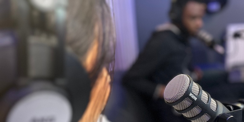 Två personer i en radiostudio, en i förgrunden med hörlurar och mikrofon, den andra suddig i bakgrunden.