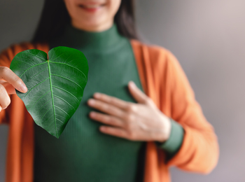 Kvinna håller ett grönt hjärtformat blad framför bröstet, klädd i grön tröja och orange kofta.