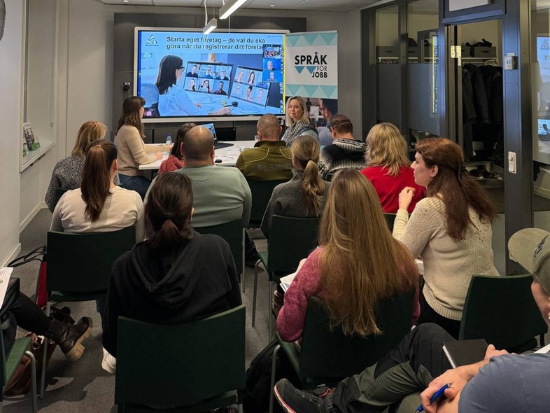 En grupp människor sitter i ett konferensrum och lyssnar på en presentation med en stor skärm längst fram.