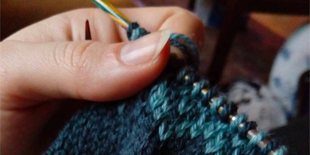 En närbild på en hand som håller i en stickning. Mönstret är mörkblått och har en ljusblå kant högst upp.