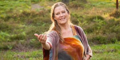 Kvinna i färgglad klänning står på en äng och sträcker ut handen, omgiven av grön natur.