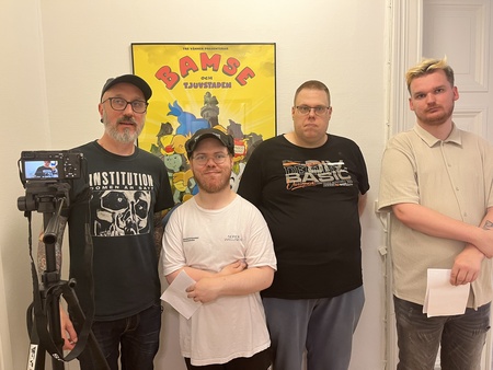 Fyra personer står framför en affisch med texten 'Bamse och tjuvstaden' i ett rum, en kamera på stativ syns till vänster.