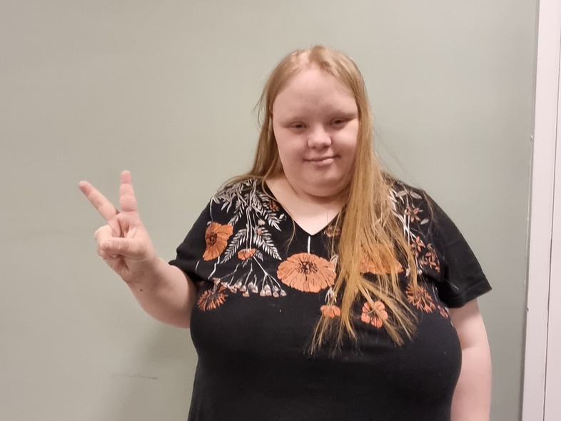 Person med långt hår och svart klänning med blommönster gör ett V-tecken med handen mot en ljus vägg.