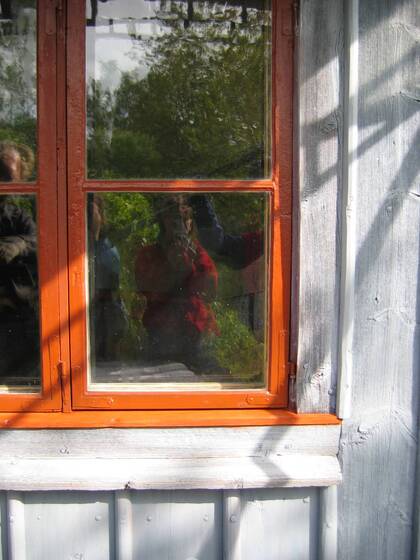 Fönster med röd ram speglar personer och grönska utanför, på en grå trävägg i solljus.