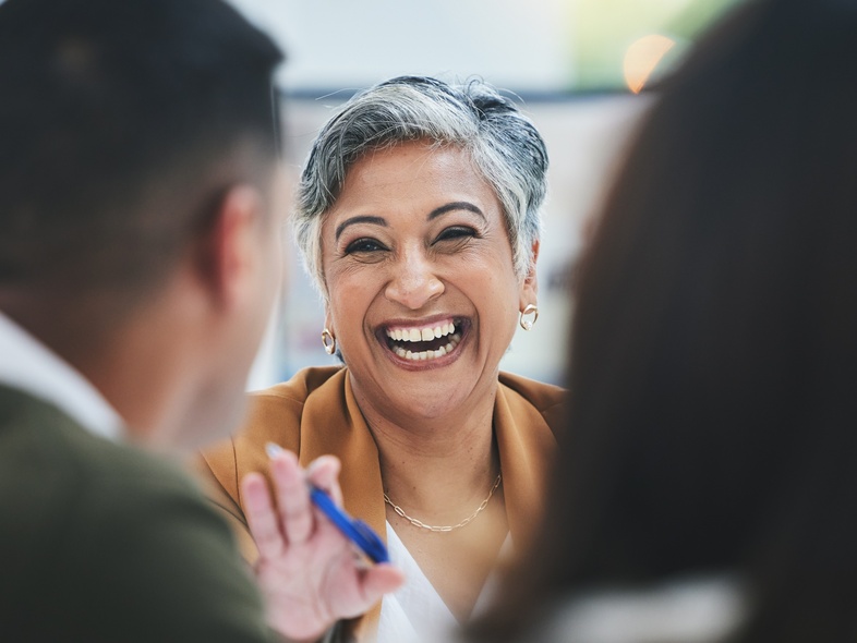 En leende kvinna med kort grått hår sitter vid ett bord och samtalar med två andra personer.