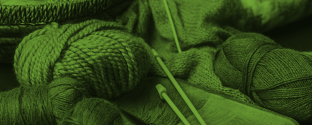 Garnnystan, stickor och sax på en bricka, med en stickad tröja i bakgrunden. Bilden har grön ton.