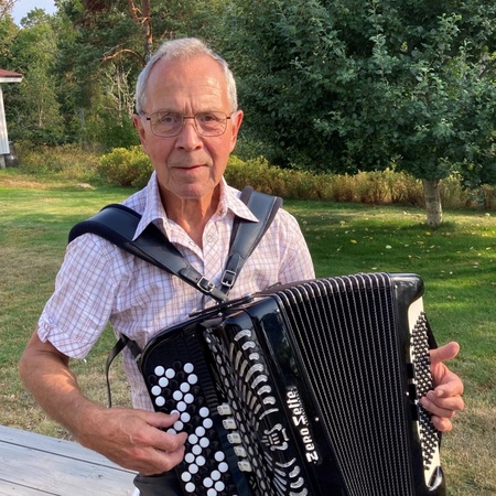 Äldre man spelar dragspel utomhus på en altan med träd och gräs i bakgrunden.