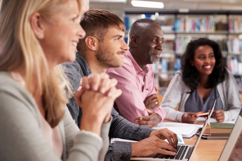 Fyra vuxna personer sitter vid ett bord i ett bibliotek och arbetar tillsammans med datorer och anteckningar.