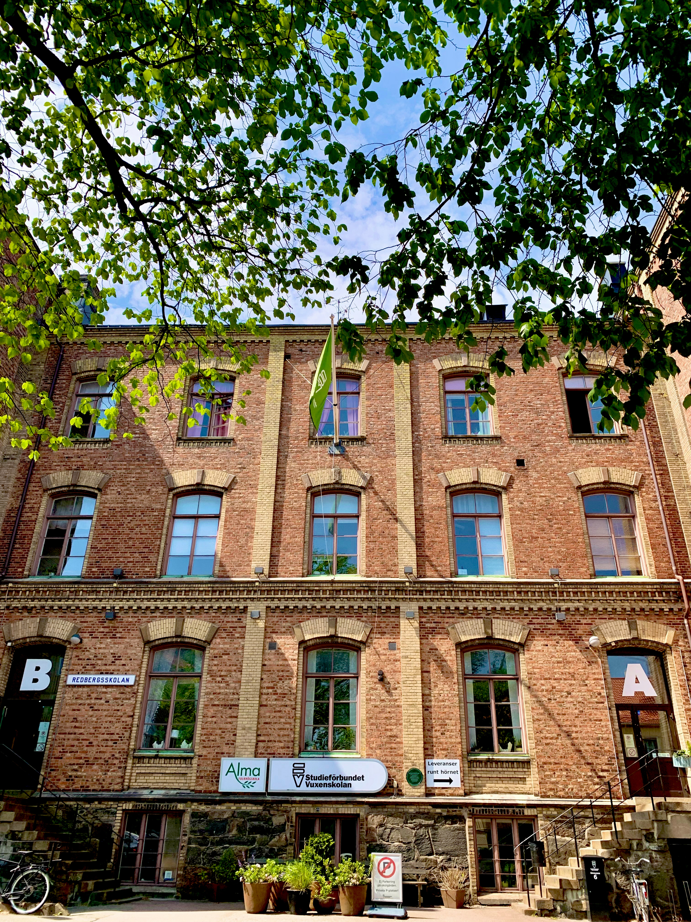 Tegelfasad på en äldre byggnad med skyltar, gröna trädgrenar i förgrunden och blå himmel ovanför.