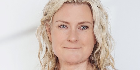 Kvinna med blont, lockigt hår i ljusblå blus står mot vit bakgrund.