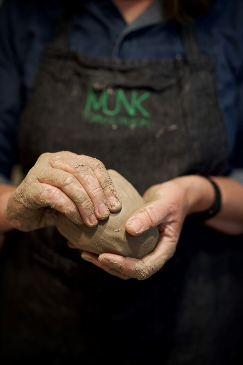 En person formar lera med händerna, iförd förkläde med texten 'MUNK'.
