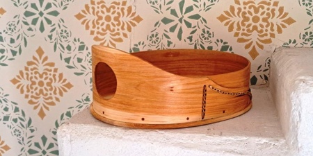 En oval svepask i trä med flätad detalj står på en vit trappa mot en vägg med grönt och gult mönster.