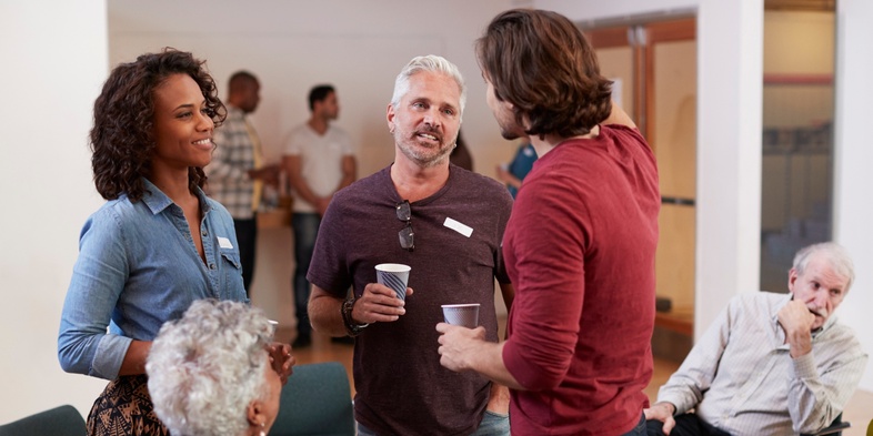 En grupp vuxna samtalar och dricker kaffe i en ljus samlingslokal med stolar i en cirkel.