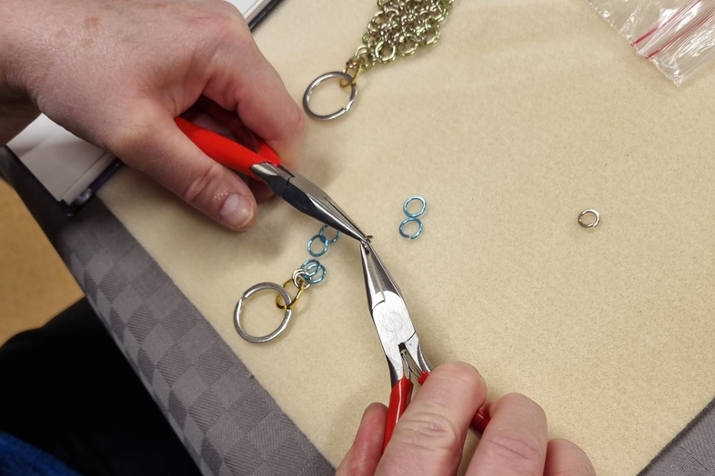 En person använder två tänger för att arbeta med små metallringar och skapa smycken på ett bord.