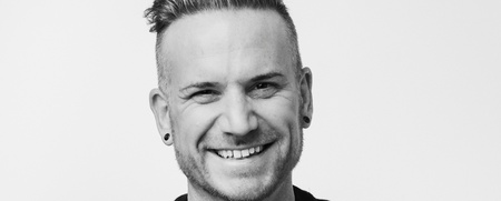 Leende man med tatuerad underarm, kort hår och svart tröja står med armarna i kors mot vit bakgrund.