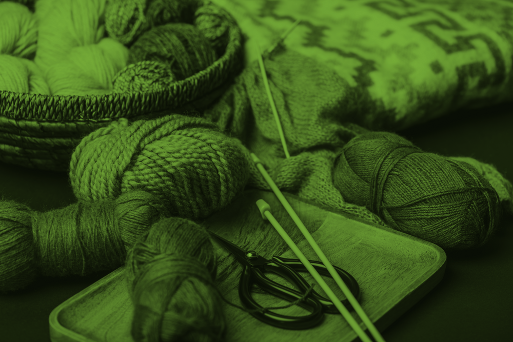 Garnnystan, stickor och sax på en bricka, med en stickad tröja i bakgrunden. Bilden har grön ton.