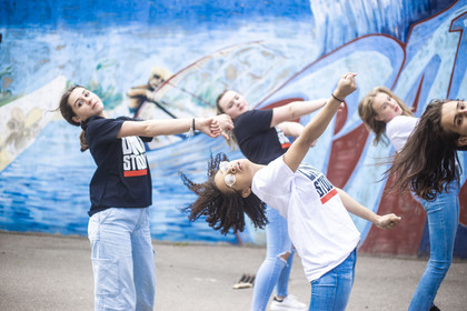 Fyra unga personer dansar energiskt framför en blå graffitivägg utomhus.