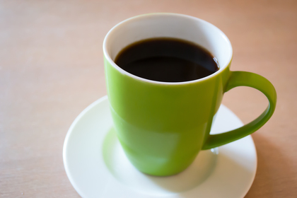 En grön kopp med svart kaffe står på ett vitt fat på ett ljust bord.