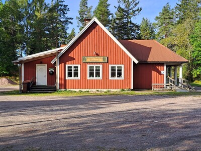 Röd träbyggnad med vita knutar och skylt, omgiven av träd och grusplan i soligt väder.