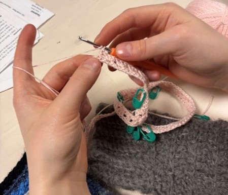 Två händer håller i ett virkningsprojekt med ljusrosa garn och gröna markörer, virknål syns tydligt.