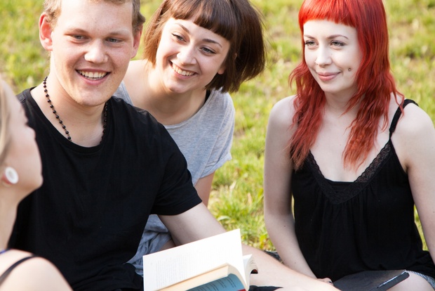 Fyra unga vuxna sitter på gräset och pratar och skrattar tillsammans utomhus.