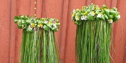 Två hängande dekorationer av långt grönt gräs och små blommor mot en röd trävägg.
