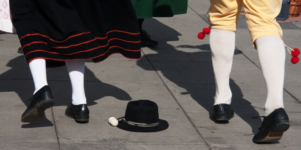Två personer i traditionella folkdräkter dansar på en stenlagd yta, en svart hatt ligger på marken.