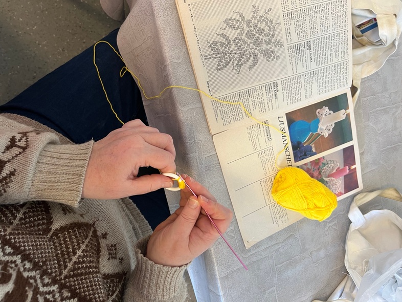Person virkar med gult garn och virknål, bredvid ett mönster i en handarbetstidning.