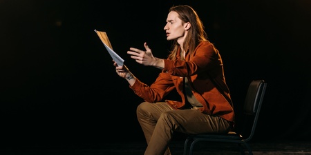 En person sitter på en stol på en scen och gestikulerar med ett manus i handen under en teaterövning.