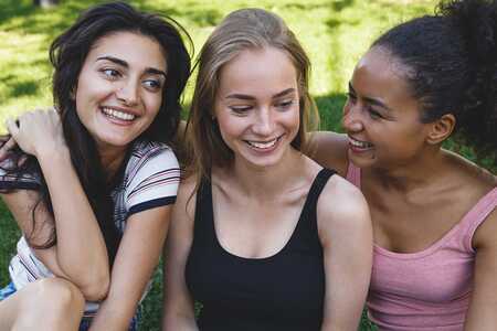 Tre leende tonåringar sitter tätt tillsammans utomhus på gräset.