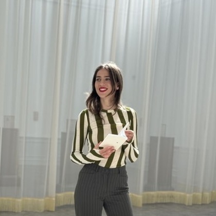 Person i randig tröja och svarta byxor står i ett ljust rum med gardiner och håller ett papper.