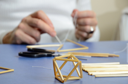 En person bygger en geometrisk form av sugrör och tråd på ett bord, med sax och fler sugrör bredvid.