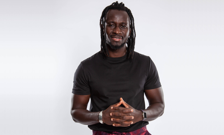 Man med mörk hy och långt flätat hår står mot vit bakgrund, bär svart t-shirt och ler lätt.