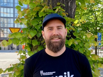 Man med skägg och keps står framför ett träd på en solig gata, bär svart t-shirt med texten 'musik'.