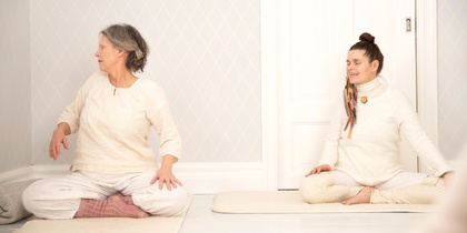 Två personer sitter på yogamattor i ett ljust rum och gör en sittande vridning.