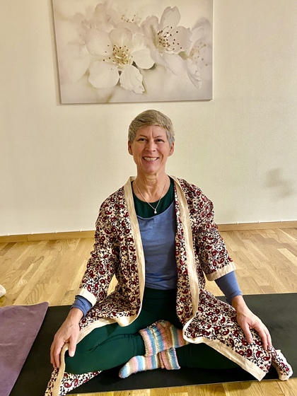 Äldre kvinna sitter leende i yogaposition på matta, iförd färgglada kläder, i ljust rum med blomkonst på väggen.