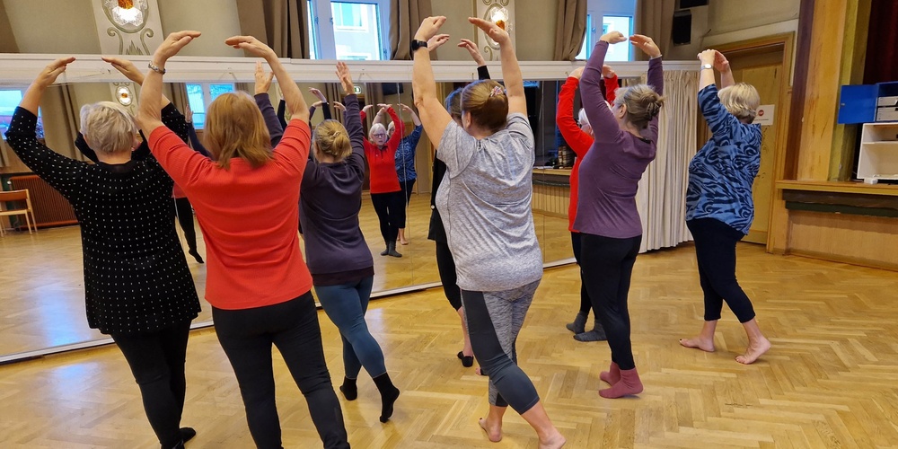 En grupp äldre kvinnor övar balett tillsammans i en spegelsal, alla står med armarna uppsträckta.