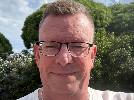 Person i vit t-shirt utomhus med buskar och molnig himmel i bakgrunden. 