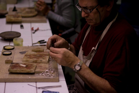 En person med glasögon och förkläde arbetar med smyckestillverkning vid ett bord med verktyg och material.