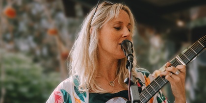 Kvinna i färgglad klänning spelar gitarr och sjunger vid en mikrofon på en utomhusscen.