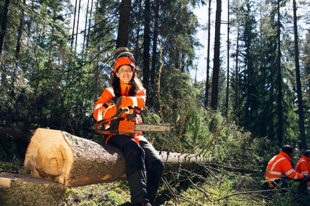 En person i orange skyddskläder sitter på en fälld trädstam i en skog med en motorsåg i händerna.