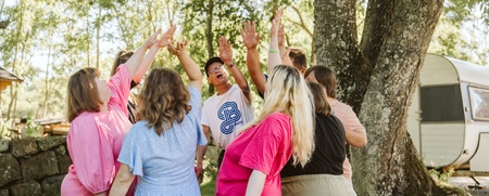 En grupp människor står i en ring utomhus och sträcker upp händerna mot mitten under ett träd.
