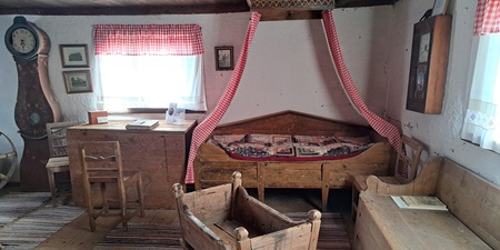 Ett gammaldags sovrum med trämöbler, vagga, säng med rödvit sänghimmel och randiga mattor på golvet.