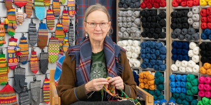 En kvinna sitter och stickar i en garnbutik omgiven av färgglada garnnystan och stickade vantar på väggen.