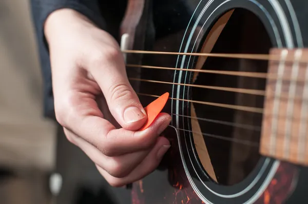 En hand spelar på en akustisk gitarr med ett orange plektrum.
