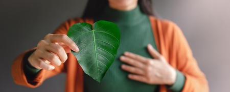 Kvinna håller ett grönt hjärtformat blad framför bröstet, klädd i grön tröja och orange kofta.