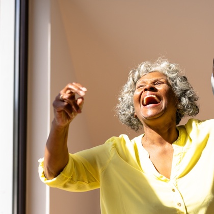 Äldre kvinna i gul skjorta skrattar och dansar med en man i ett soligt rum.