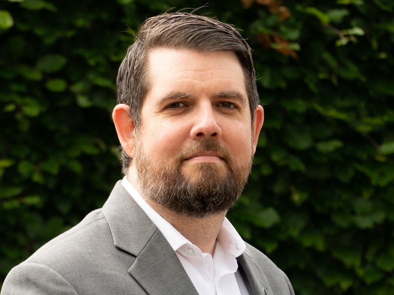 Man med skägg i grå kostym står utomhus framför en grön häck.